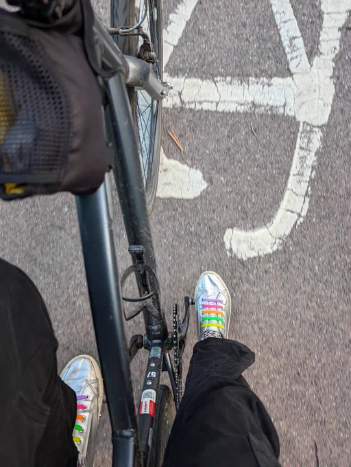 Photo of view down towards my bike pedals whilst cycling. Dark grey bicycle frame and front wheel in view plus silver shoes with rainbow laces and the cycle path beneath shows a section of a bike lane symbol