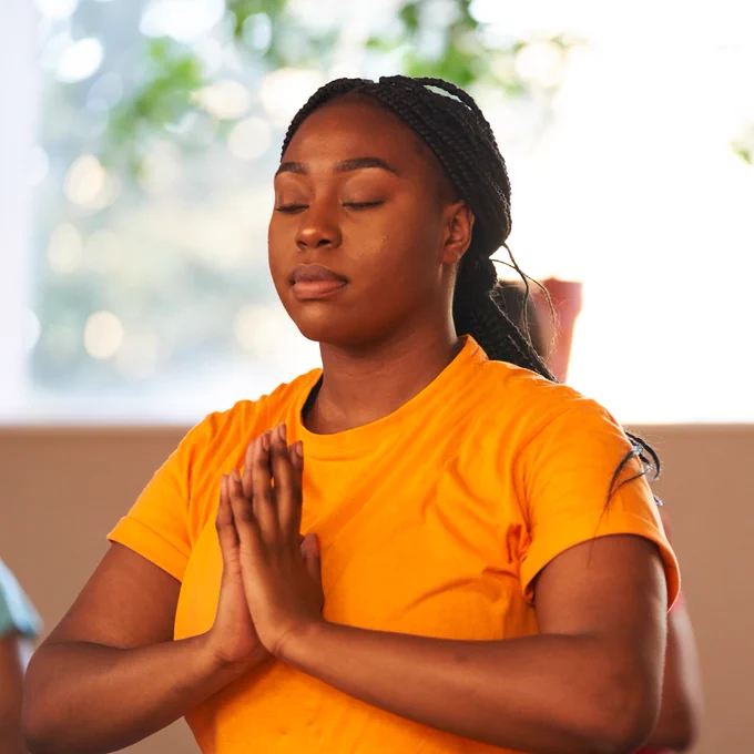 Person in a yellow shirt practicing meditation with hands pressed together, eyes closed, in a serene indoor setting.