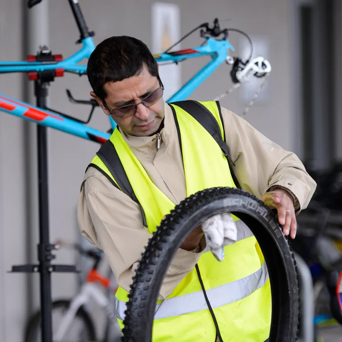 A volunteer in a high-visibility vest inspects a bicycle tire in a workshop with bike racks in the background.