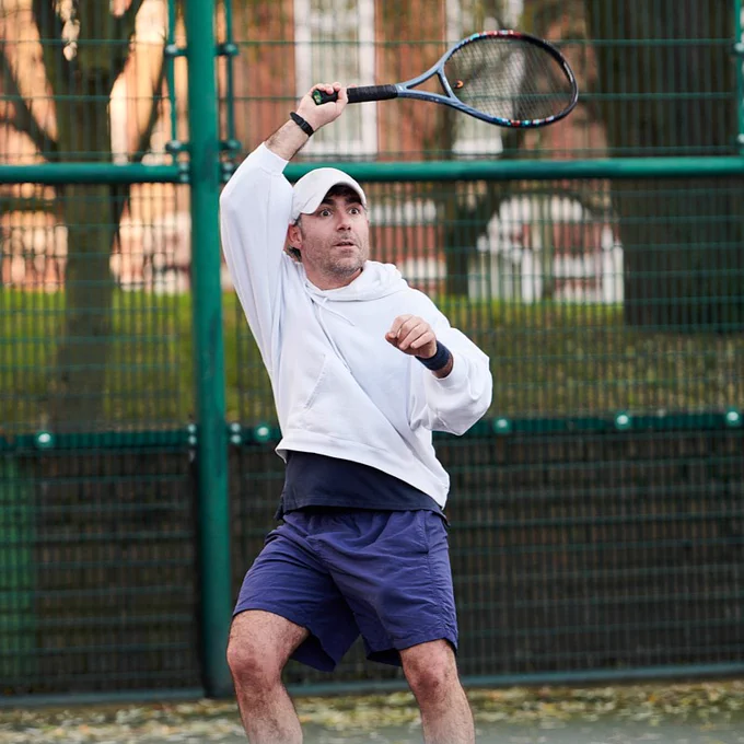 Person playing tennis, dressed in a white long-sleeve shirt and blue shorts, swinging a racquet overhead on a court enclosed by a green fence.