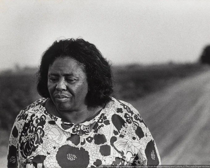 A gelatin silver print of Fannie Lou Hamer wearing a floral print top. She is standing on a dirt road. 
