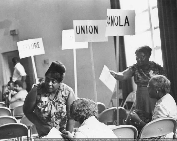 A black-and-white photograph of Fannie Lou Hamer during the Mississippi Freedom Democratic Party Convention (1964). Three protest signs in the distance read [PANOLA] [EXPLORE] [UNION].