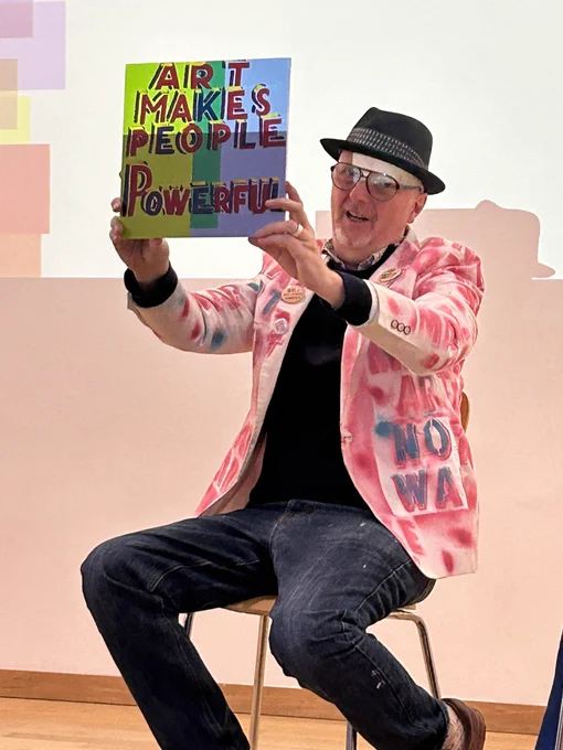 A man, seated, holds up a painting: it reads ‘Art makes people powerful’.