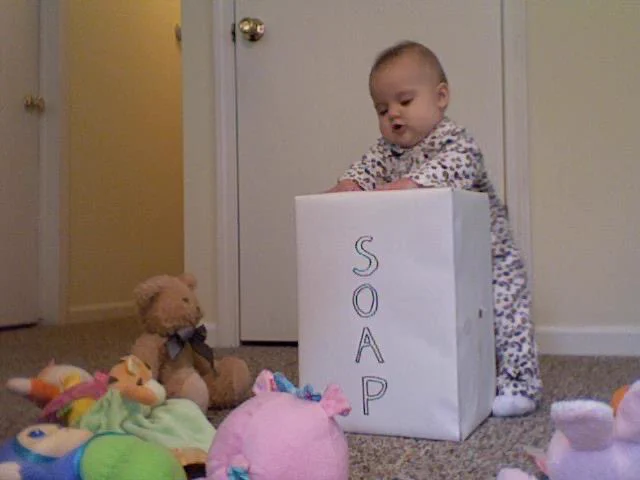 A baby in patterned jammies stands at a box like it’s a lectern, surrounded by an audience of soft toys
