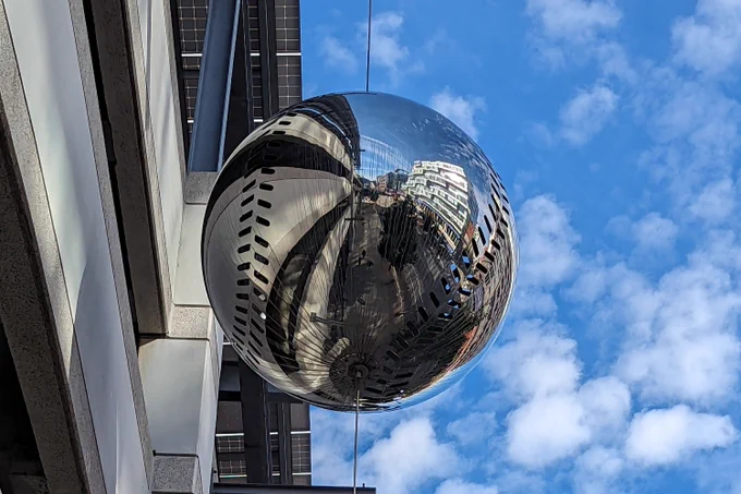 Reflective spherical sculpture hanging between buildings, distorting surrounding cityscape and sky in its mirrored surface.