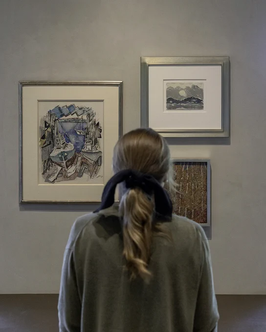 Photograph of a person from behind with a bow in their hair. The person is looking at three paintings ( Eastport Docks, Maine Coast, by John Marin; Untitled (Mountain and Sun) by Arthur Dove; Untitled by Marsden Hartly) on a wall.