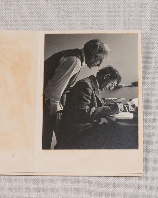 Photograph of Alfred Stieglitz in wearing glasses, a light shirt and waistcoat bending over and looking over the shoulder of John Marin who wears glasses, a jacket, and is writing at a desk.