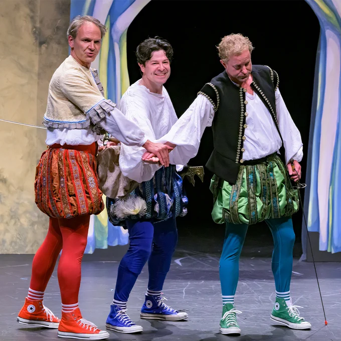 Three men wearing Shakespeare costumes place their hands together.