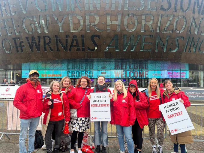 A group of people wearing Crisis branded clothes in front of the Wales Millenium Centre. They are holding signs which read ‘United against homelessness’ and ‘hanner ffordd gartref’