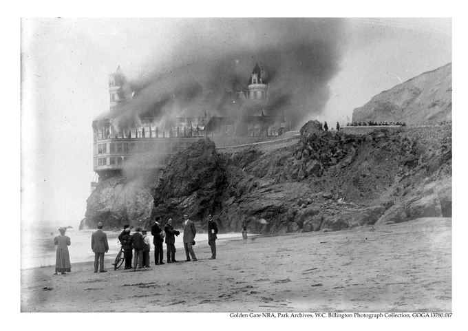 People stand on a beach watching as smoke billows out of a large building.
