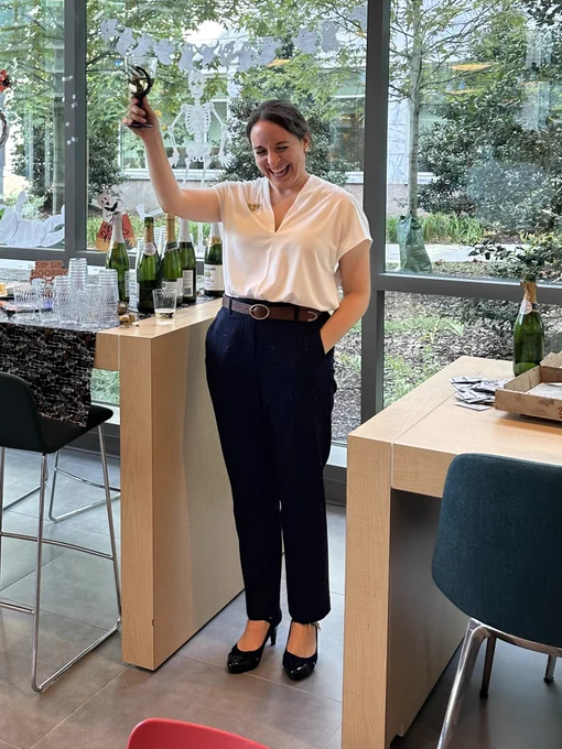 Brooke holds up a glass to toast, dressed in professional slacks, a blouse, and heels. She is laughing and surrounded by Halloween decorations and party food.