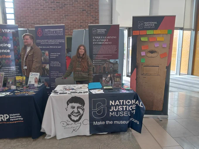 A table with organisation banners on. Has various leaflets and items on the table. A young woman stands behind the table looking at the camera.