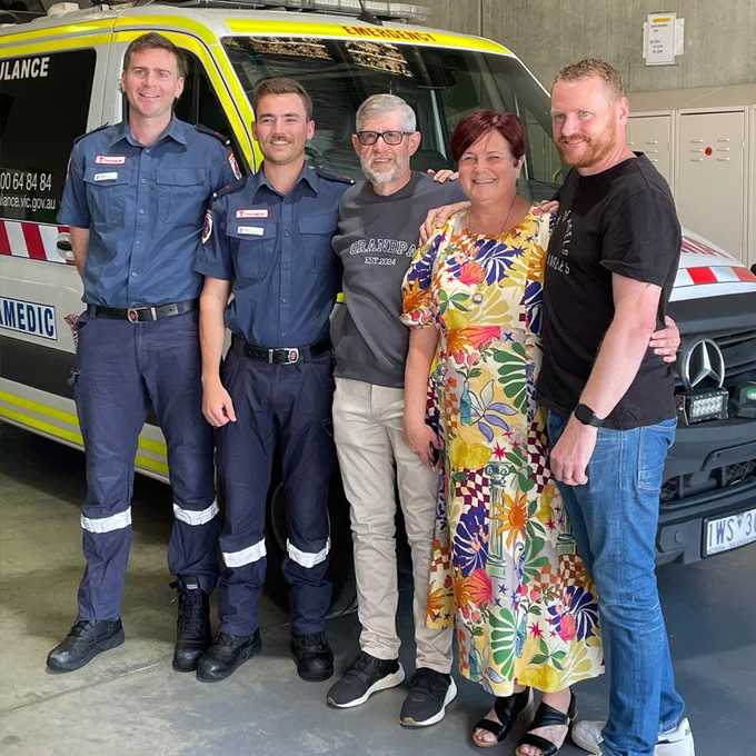 Five adults, including two in paramedic uniform, standing in front of an ambulance vehicle in a garage.