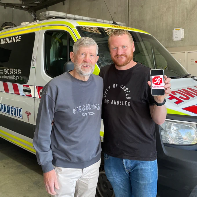 Two individuals smiling in front of an ambulance vehicle, one holding a smartphone displaying the GoodSAM app. 