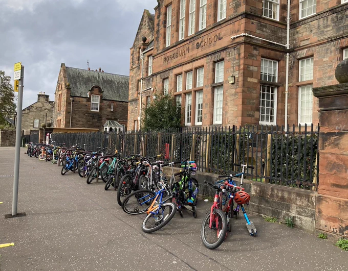 Over twenty children's cycles and a smattering of kick-scooters are locked to black sheffield stand cycle racks outside of the red-bricked Roseburn Primary School - their frames a riot of colour on an otherwise overcast day.