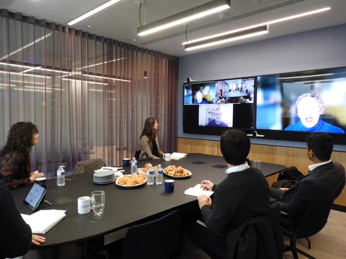A group of us around the table in the Zoom offices. Some are in the physical room and others on Zoom. We are looking at the screen. There are coffee cups and croissants on the table.