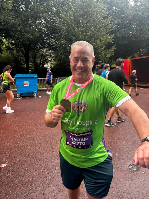 A man in a green charity running top and shorts smiles as he holds his race medal