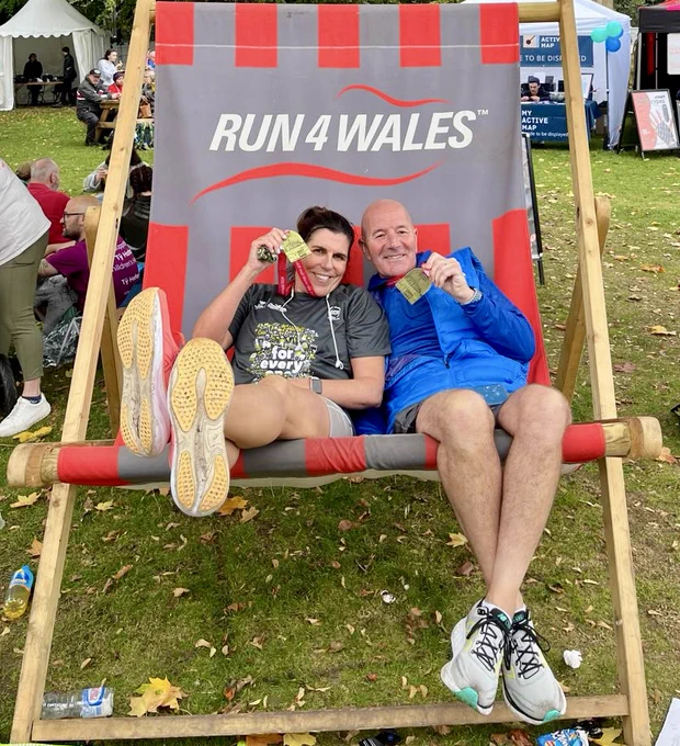 A woman and man sit on a giant grey and red striped deckchair with their race medals.