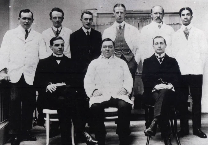 A black and white formal photograph of a group of men in dated suits or white lab coats staring into the camera