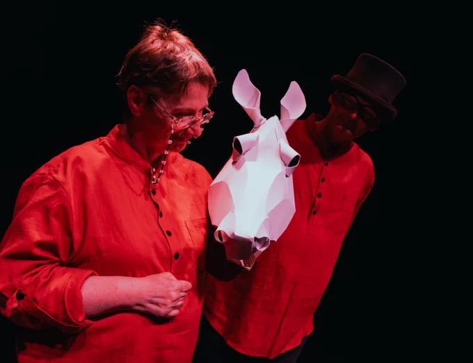 Two actors, one wearing a top hat and both looking at a white paper sculpted horse head held by the other actor. 