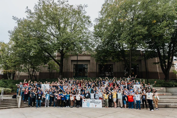 A group photo of all DjangoCon US 2024 in-person attendees outdoors on steps with their hands outstretched. Behind them are trees and a cement building.