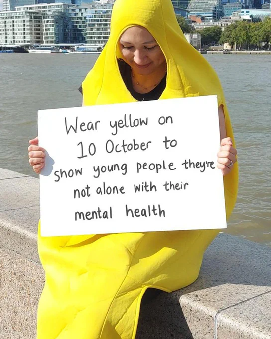 A women in a banana suit, sitting on the bank of the Thames with the city in the background, holding up a sign that says 'Wear yellow on 10 October to show young people they're not alone with their mental health'. 