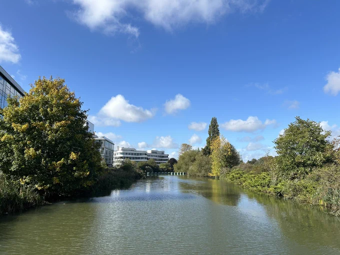 Photograph shows the River Nene taken from the bridge on Nunn Mills Road, Northampton. The river is in the centre of the image with the tree and bush lined banks on the left and right sides. On the left behind the trees and bushes is Lancaster House. To the rear left and rear centre of the photograph are the Learning Hub and the Creative Hub of the University of Northampton respectively.