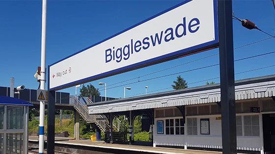 The Biggleswade station sign with platforms in the background on a lovely sunny day.