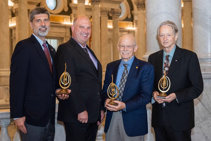Four men in formal attire stand in a grand marble hall, each holding the Golden Goose Award trophy – a golden spiral design resembling a goose. From left to right, we have Karl Rumelhart and Peter Rumelhart, James McClelland, Geoffrey Hinton. They are smiling and posing together. The background features classical architecture with columns and arches, adding to the formal atmosphere of the event. (photo by Linnea Farnsworth)