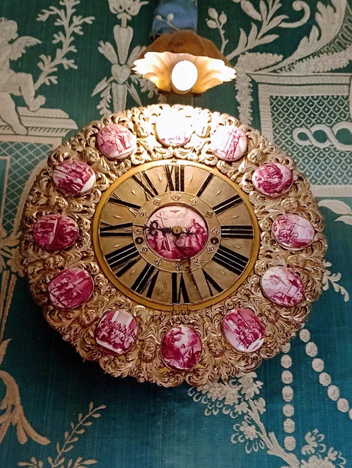 An ornate wall clock with a gold coloured face, surrounded and centered with small rose coloured cameos of what might be biblical scenes