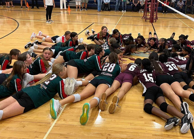 Members of two opposing volleyball teams laying on the court in a circle.