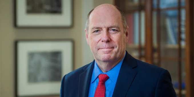 Portrait photo of RPI President Martin A. Schmidt wearing a blue suit and red tie, standing in an office with framed pictures and a bookcase in the background.