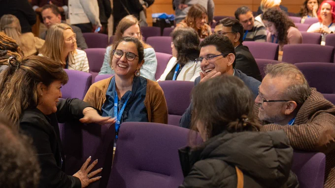 2024 Symposium attendees smiling as they have a conversation while sitting in purple chairs surrounded by other participants. 