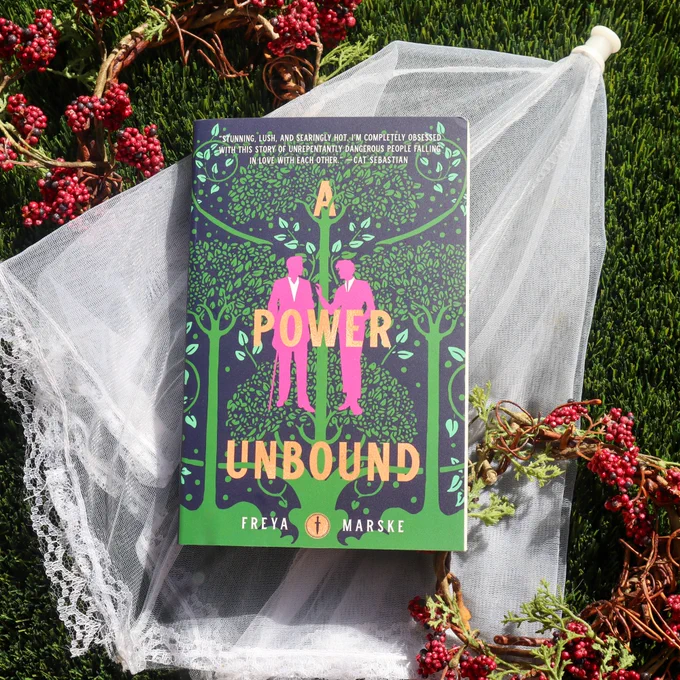 A paperback copy of A Power Unbound by Freya Marske is laying on a white lace fabric and red berries surround the book.