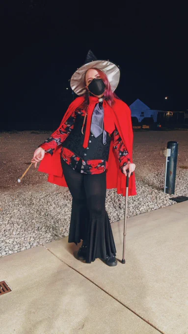 Full body photo of Jules dressed as a wizard. They are white with medium length purple hair with long braids on either side and they're wearing black dramatically flared pants, a black red and silver long sleeve button down with a rose print all over it, a black corset with a subtle baroque pattern, a red cloak, a big red and silver bowtie with a sparkly star in the center, and a black and silver pointy hat. They are standing with a silver and black cane in their left hand and a wand with a moonstone at the end in their right hand, lifting it slightly.
