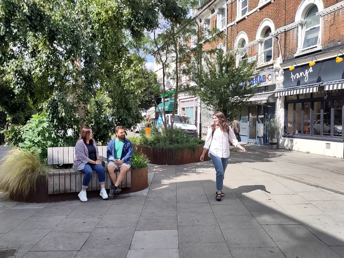 Local people enjoying a pocket park. Two people are sitting on a bench, and another is walking past
