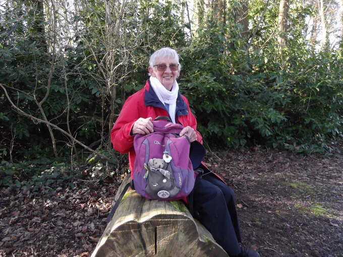 Delia is taking a rest from walking and sitting on a log with her little companion Team Together Ted, who rides in the pouch of her backpack. 