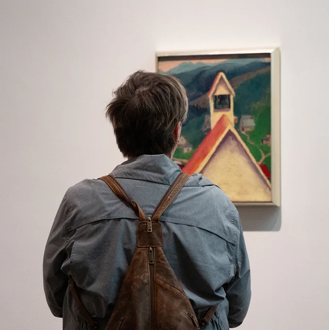 Photograph of a person taken from behind as they look at a small painting of a church steeple on a white gallery wall.