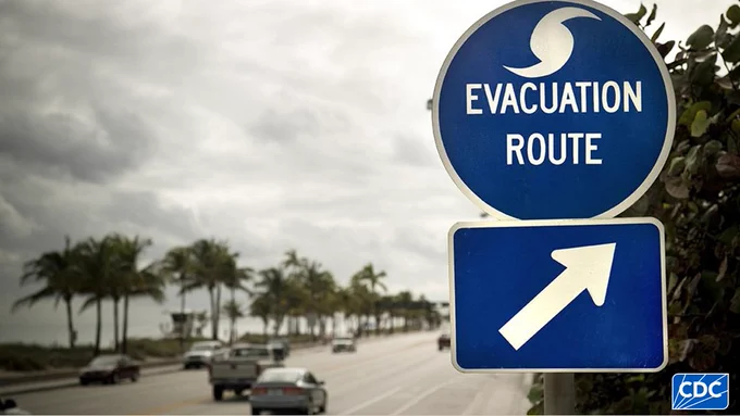 A blue sign that says “Evacuation Route” with an arrow directing people toward a safe route. Cars are driving along a road in the direction the arrow is pointing.