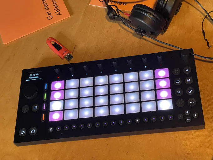 Move hardware next to a pair of headphones, a USB key, ans starter guides, showing light up 32 pad grid, encoders, buttons and display.