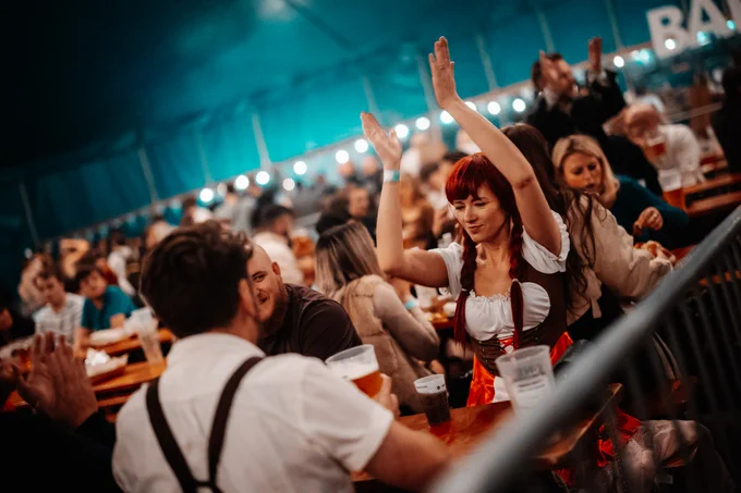 People in traditional Bavarian attire enjoying themselves at Oktoberfest, with one individual raising their arms and smiling, surrounded by others seated at long tables.