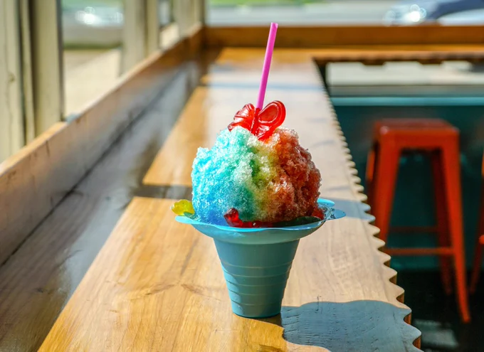 A brightly coloured rainbow slushy-ice, with a candy bow on top, in a bowl, sitting on a wooden shelf as sun shines in through the window.