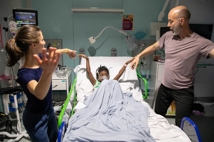 A young girl lies in a hospital bed, raising her arms while two adults, one on each side, mirror her movements playfully. The woman on the left and the man on the right extend their arms, engaging the child in a lighthearted dance. The hospital setting is visible, with medical equipment and monitors in the background. 