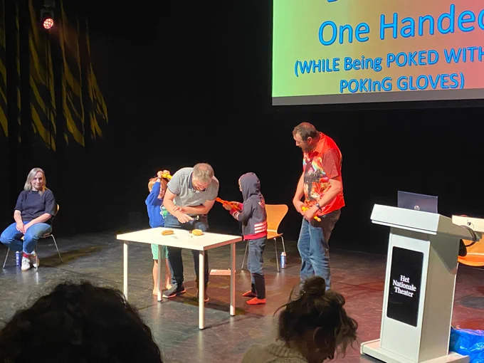 Three adults and two children are on stage in a studio space. The children are poking one of the adults in a grey t shirt with big foam fingers and laughing.  At the back of the stage there is a screen that reads: 'One handed' and to the right of the image is a lecturn.