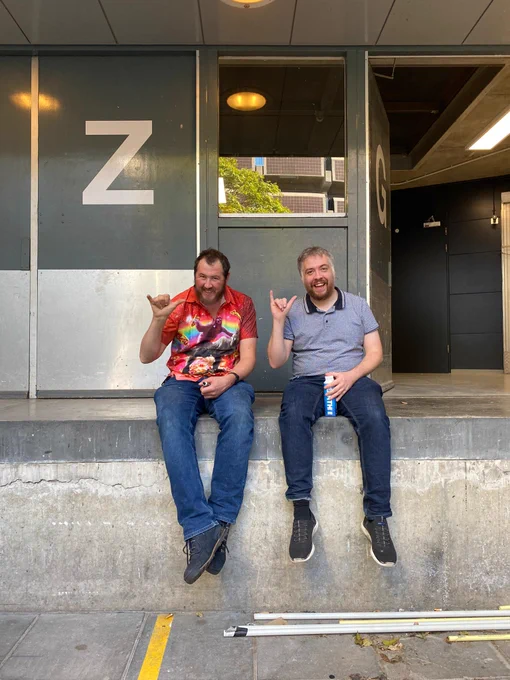 A man in a red shirt and jeans and a man in a grey t shirt and jeans are sat on a concrete wall making the 'hang loose' gesture