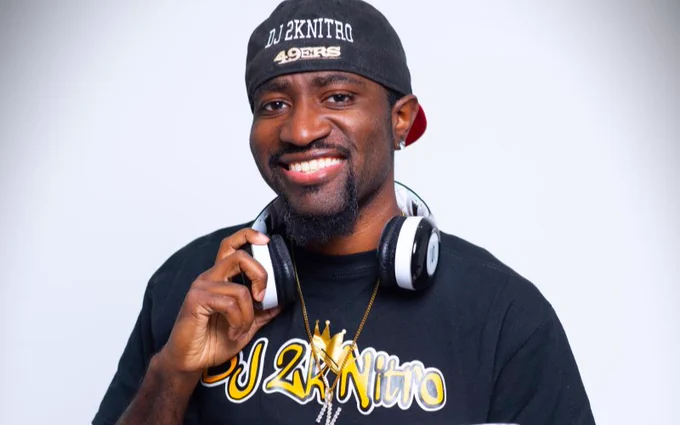 A person wearing a San Francisco 49ers cap and headphones smiles while holding a smartphone to their ear. They are dressed in a black t-shirt with visible text and are standing against a plain light background.