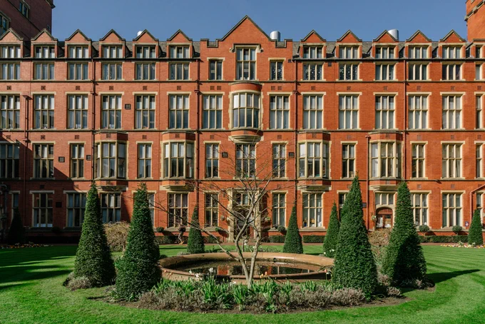 A red brick building with lots of windows. In front is a green lawn and a circular water feature surrounded by conical topiary.