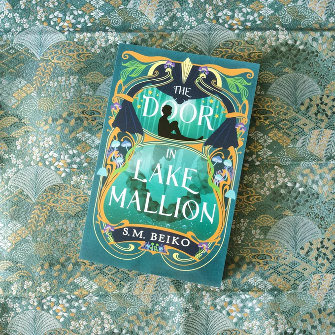 The book The Door in Lake Mallion by S.M. Beiko lays on top of teal fabric with intricate white and metallic gold patterns.