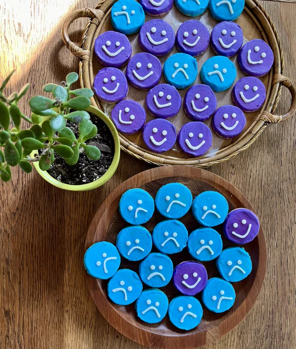 Two wooden platters w blue sad face cookies and purple happy face cookies and a jade plant in a lime green pot all on a wood table