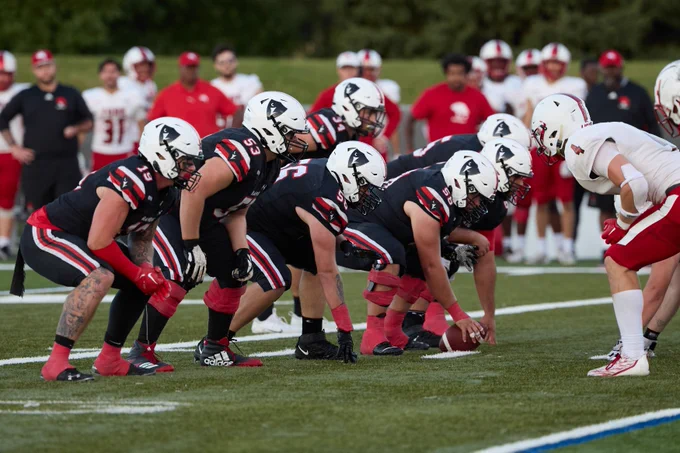 Football players from two teams, one wearing black and red uniforms with helmets featuring a hawk logo, and the other in white and red, are lined up facing each other on a football field, poised for the start of a play.
